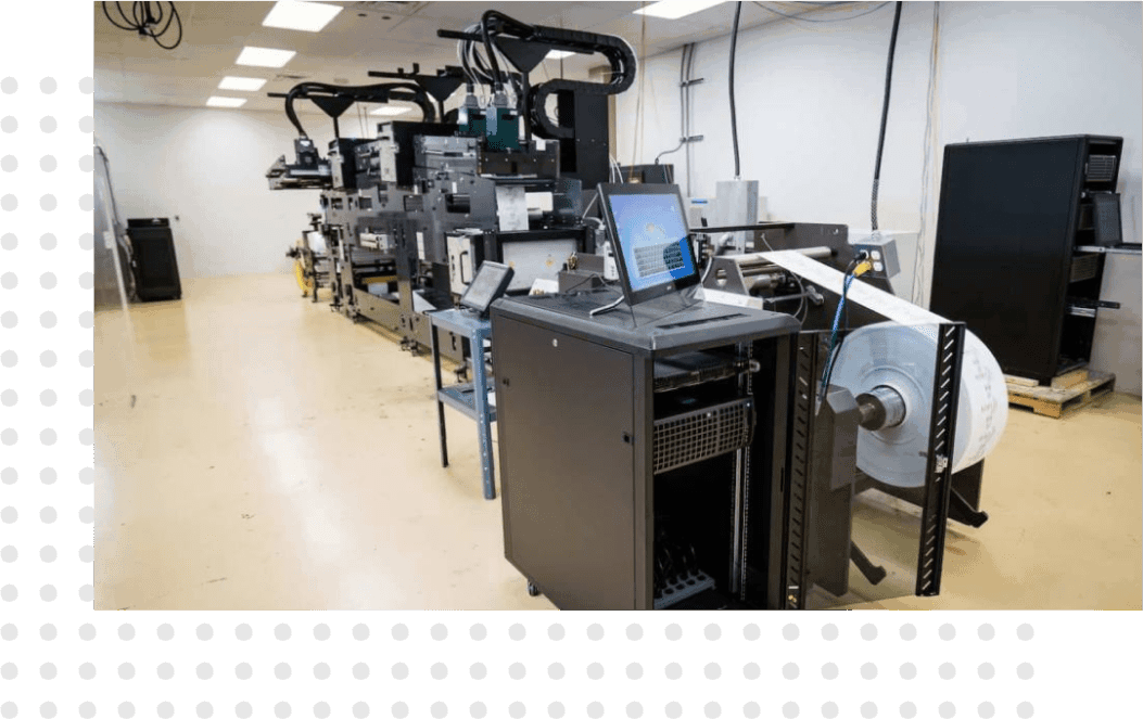 An industrial printing setup inside a well-lit room. The equipment includes large printing machines with various components and control panels.